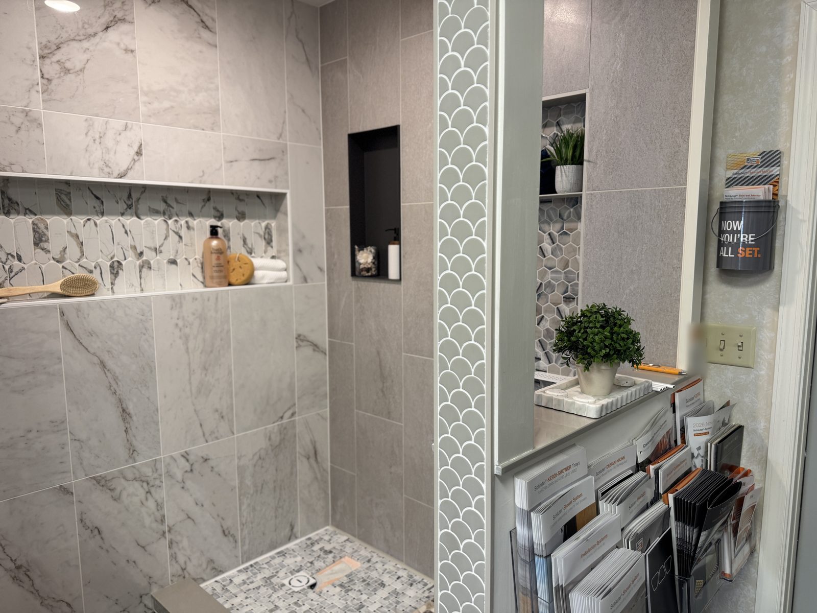 Close-up of the display shower: a marble mosaic niche, a black metal niche, a scalloped tile border, and Schluter shower system brochures